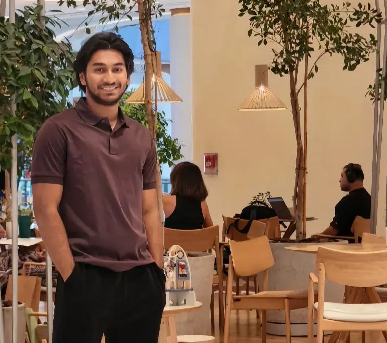 Nikesh smiling in a plum polo shirt beside indoor plants and cafe seating.
